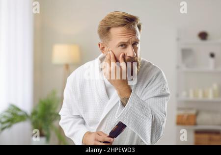 Uomo che esamina il suo volto al mattino controllando la condizione della pelle e la presenza di rughe. Foto Stock
