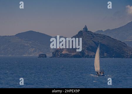 Scena del mare con una barca a vela in direzione di terra. Un vecchio faro costruito sulla cima di un'isola rocciosa si affaccia sul soggetto. Il cielo è limpido e c'è una leggera nebbia nell'aria, il che suggerisce una giornata calda e muggy. Foto Stock