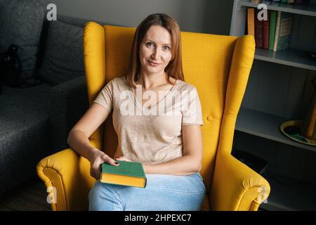 Vista ad alto angolo della bella donna bionda che riposa in una comoda sedia gialla che tiene un libro di carta in camera oscura con interni moderni Foto Stock