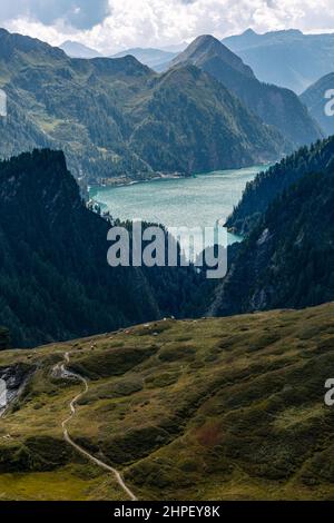 Veduta aerea di un paesaggio alpino con un sentiero che si snoda attraverso i pascoli. Sul sentiero ci sono due persone che camminano verso alcune mucche. Sullo sfondo si trova un lago di montagna, verso il quale si dirige il sentiero. Foto Stock