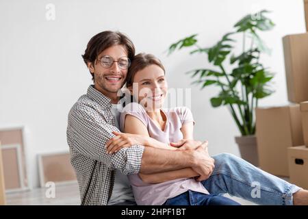 Soddisfatto giovane europeo in occhiali abbraccia signora in casual, si siede sul pavimento tra scatole di cartone Foto Stock