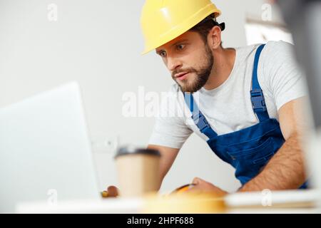 Un uomo di mano concentrato in uniforme e un elmetto utilizzando il computer portatile mentre si inizia il lavoro di ristrutturazione in interni Foto Stock
