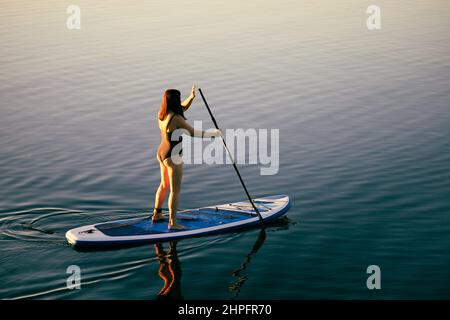 Vista dall'alto su una donna rilassata di mezza età su sup board canottaggio con OAR su acqua di lago calma. Stile di vita attivo. Attività di svago per le persone di Foto Stock