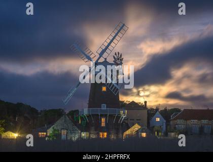 Una luna piena che si innalza attraverso le folte nuvole dietro il mulino a vento a Cley accanto al mare, lungo il fiume Glaven sulla costa di Norfolk, Inghilterra, Regno Unito Foto Stock