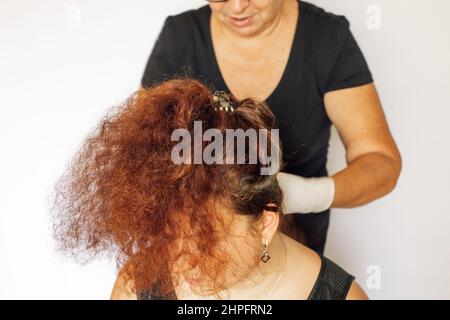 Donna anziana indossando guanti bianchi in lattice sulle mani tintura ricci capelli rigogliosi di donna di colore rosso scuro su sfondo bianco. Colorazione dei capelli a casa Foto Stock