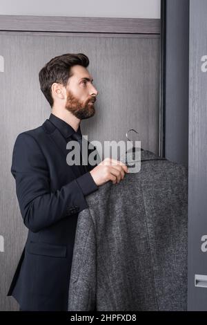 vista laterale dell'uomo in bearded nel gruccia di tenuta del vestito con cappotto vicino all'armadio nella camera dell'hotel Foto Stock