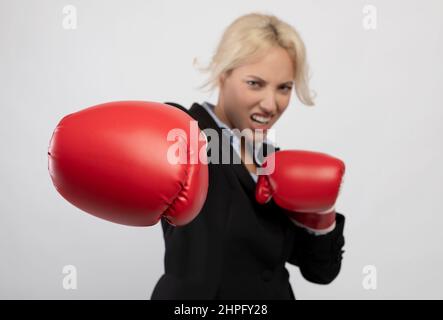 Giovane donna d'affari boxe che punisce verso la macchina fotografica indossando guanti da boxe rossi, fuoco selettivo, primo piano, sfondo chiaro Foto Stock