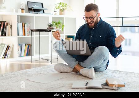 Uomo gioioso usando il laptop urla e gesturando Sì seduto in casa Foto Stock