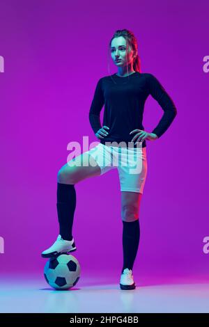 Ritratto di bella ragazza, professionista giocatore di calcio femminile in posa con palla di calcio isolato su sfondo viola studio in luce al neon. Sport Foto Stock