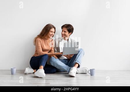 Soddisfatto giovane coppia caucasica guardare laptop progettazione di nuovi interni, sedersi a pavimento in stanza vuota su sfondo bianco parete. Comprando bene immobile, grande Foto Stock