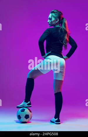 Ritratto di bella ragazza, professionista giocatore di calcio femminile in posa con palla di calcio isolato su sfondo viola studio in luce al neon. Sport Foto Stock