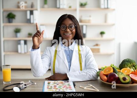 Sorpreso nutrizionista africano americano seduta al posto di lavoro e alzando il dito in su, avendo idea mentre scrive il programma di dieta Foto Stock