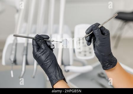 Primo piano delle mani del medico femminile in guanti neri che tengono gli strumenti dentali - sonda e specchio. Concetto di cura e trattamento dei denti, clinica professionale di stomatologia. Foto Stock