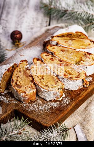 Stollen di Natale su un tagliere. Sfondo bianco tavolo in legno. Tradizionale dolce natalizio con marzapane, noci, uvetta e frutta secca. Verticale Foto Stock