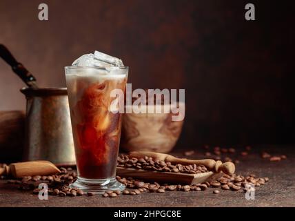 Il caffè del ghiaccio con il panna che viene versato in esso che mostra la consistenza e l'aspetto rinfrescante della bevanda. Bicchiere e chicchi di caffè surgelati su un vecchio tavolo marrone wi Foto Stock