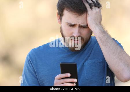 Ritratto della vista frontale di un uomo preoccupato che controlla lo smartphone all'aperto Foto Stock
