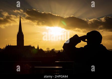 SALISBURY, REGNO UNITO. Un uomo guarda il sole tramontare sopra la Cattedrale di Salisbury in condizioni molto più calme dopo giorni di forti venti e forti piogge nel Wiltshire, Regno Unito. Data: 21/02/2022. Credit: Matthew Lofthouse/Alamy Live News Foto Stock