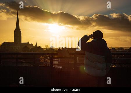 SALISBURY, REGNO UNITO. Un uomo guarda il sole tramontare sopra la Cattedrale di Salisbury in condizioni molto più calme dopo giorni di forti venti e forti piogge nel Wiltshire, Regno Unito. Data: 21/02/2022. Credit: Matthew Lofthouse/Alamy Live News Foto Stock