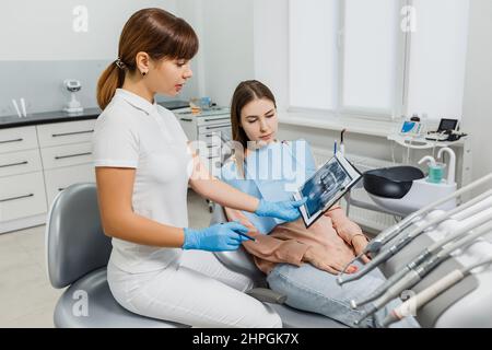 Dentista amichevole con tablet e immagini radiografiche che spiegano il trattamento al paziente durante l'esame dei denti. Moderna clinica dentale. Concetto di cura orale professionale, consultazione in studio di stomatologia. Foto Stock