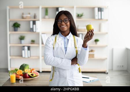 Allegro consulente di nutrizione femminile afroamericana con mela e metro a nastro che lavora in clinica e sorridendo alla macchina fotografica. Promozione speciale perdita di peso Foto Stock