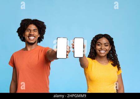 Uomo e donna neri sorridenti e millennial in t-shirt colorate che mostrano moderni telefoni cellulari con schermi bianchi vuoti, consigliando il nuovo divertente mobi Foto Stock