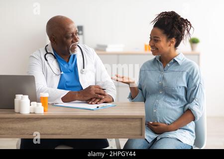 Concetto di cura prenatale. Donna incinta di colore nero felice seduta in clinica o ospedale, avendo consolazione con ginecologo maschio maturo, abbracciando il ventre. Lui Foto Stock