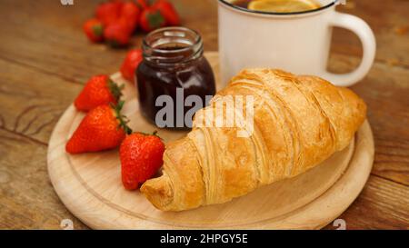 Pausa tè snack. Tè al limone, croissant, vasetto di marmellata e fragole fresche su sfondo di legno. Colazione dolce e gustosa. Concentratevi sul croissant appena sfornato Foto Stock