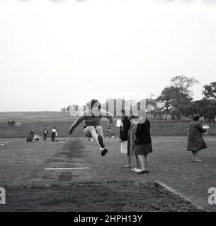 1960s, storica, una giovane atleta femminile a mezz'aria che fa il salto lungo, Fife, Scozia, Regno Unito. Velocità, forza, agilità e determinazione sono tutti necessari in questo grande evento su pista e campo, dove l'obiettivo è quello di saltare il più possibile in un sandpit da un punto di decollo della corsa. Evento maschile nelle Olimpiadi moderne originali del 1896, le donne hanno partecipato per la prima volta al salto olimpico lungo nel 1948 a Londra. Foto Stock