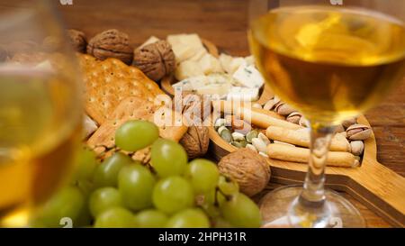 Spuntini per vino. Piatto di formaggi. Formaggio, noci, uva, cracker su sfondo ligneo. Primo piano sfocato con due bicchieri di vino bianco. Foto Stock