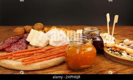 Piatto di carne e formaggio per antipasti. Vasetti con marmellata e miele. Salsicce, formaggi, cracker, noci su un piatto di legno e sfondo. Foto Stock