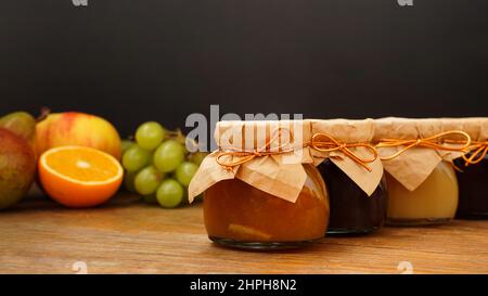 Marmellata fatta in casa, fuoco selettivo. Vasetti con confit. Dolce dessert. Frutti sfocati sullo sfondo. Uva, arancia, mela. Marmellata fatta in casa Foto Stock
