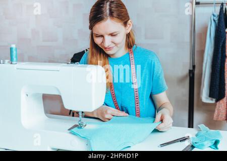 Un sarto femminile fa un modello . Giovane donna sorridente stilista che lavora sulla scrivania su una macchina da cucire. Dressmaker che fa un indumento sul posto di lavoro Foto Stock