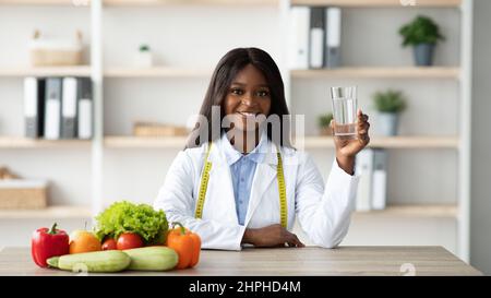 Giovane nutrizionista afroamericana che tiene un bicchiere d'acqua mentre si siede a tavola con frutta e verdura Foto Stock