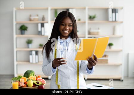 Positivo dietista nero con taccuino e bicchiere d'acqua, in piedi alla clinica e sorridendo alla macchina fotografica, spazio libero Foto Stock