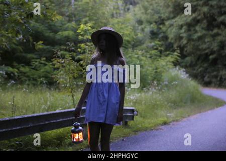 Piccola ragazza carina in un cappello di paglia. Passeggiata serale nella foresta con un'antica lanterna nelle sue mani. Estate, luce del sole, ritratto, caldo, foresta. Foto Stock