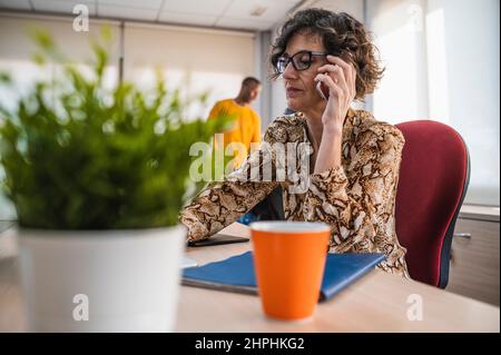 Donna anziana che parla al telefono mentre lavora in ufficio. Foto Stock