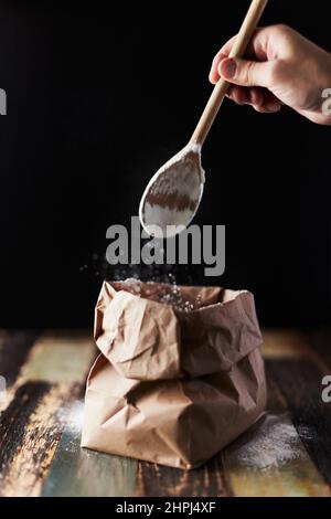 Farina di grano in un pacchetto di mestiere. Movimento di congelamento, cucchiaio sfondo scuro, spolverare la farina. Vista frontale. Foto Stock