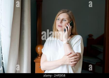 Donna adulta felice che parla al telefono mentre si alza alla finestra contro le scale di sfondo nel soggiorno. La donna caucasica comunica utilizzando la connessione cellulare in abiti casual, guarda verso la finestra Foto Stock