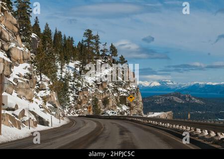 vista panoramica su un'ampia strada costeggiata da montagne rocciose innevate con pini da un lato e profonde valli dall'altro, sfondo di rema Foto Stock