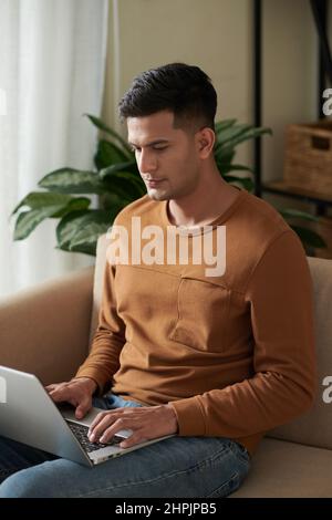 Giovane uomo seduto sul divano nel soggiorno con un computer portatile e concentrato sul suo lavoro online su un computer portatile Foto Stock