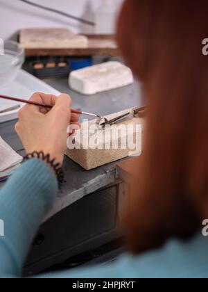 una donna che lavora nel suo laboratorio artigianale di gioielleria utilizzando una piccola spazzola su un anello per eseguire il suo lavoro su un blocco di pietra Foto Stock