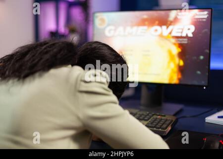 Triste perdente giocatore persona giocare spazio sparatutto video giochi perdere campionato online. Giocatore nervoso afroamericano che usa l'apparecchiatura del calcolatore RGB seduta nello studio domestico di gioco. Torneo di eSport Foto Stock