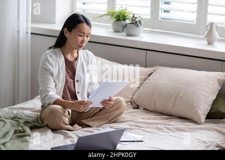 Giovane auditor freelance femminile contemporaneo che legge i documenti finanziari mentre si siede a letto di fronte al laptop e lavora da casa Foto Stock