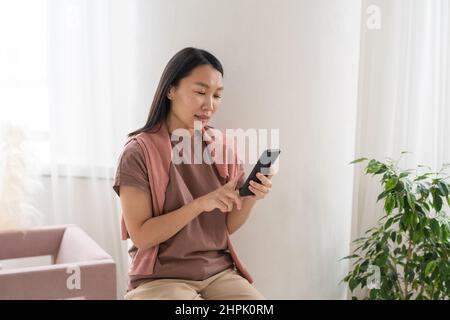 Giovane ragazza graziosa di etnia asiatica che punta allo schermo dello smartphone mentre messaging o scorre attraverso i prodotti in linea nel paese Foto Stock