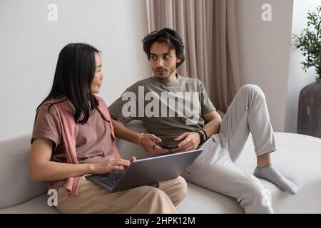 Giovane uomo d'affari fiducioso con un laptop che parla con un dipendente asiatico, sia seduto sul divano che discutendo dati online Foto Stock