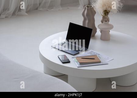 Posto di lavoro di freelance contemporaneo con gadget mobili, vasi fatti a mano, notebook e documenti finanziari su tavola rotonda bianca Foto Stock