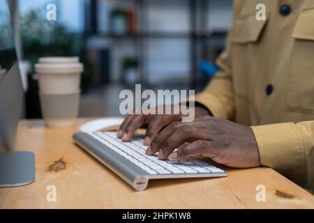 Mani di un giovane uomo d'affari afroamericano premendo i tasti della tastiera del computer mentre si lavora in internet da un tavolo di legno Foto Stock