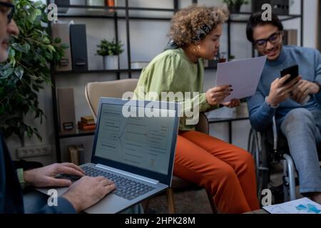 Economista maturo che analizza i dati sullo schermo del laptop contro due giovani colleghi interculturali che discutono di documento finanziario Foto Stock