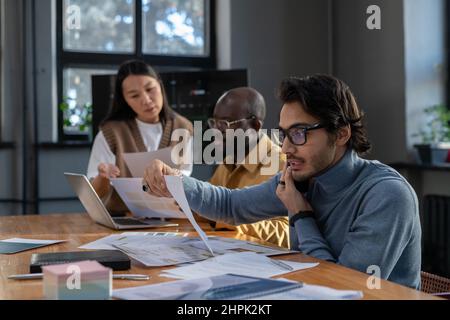 Giovane economista occupato con telefono cellulare per orecchio guardando attraverso i dati nel documento finanziario contro due colleghi che discutono di documenti Foto Stock