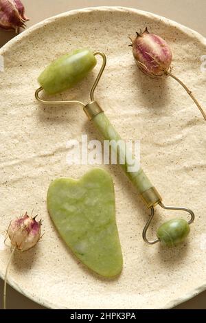 Rullo per il viso Jade gemstone e gua sha per il massaggio del viso su piastra di pietra con fiori secchi. Concetto di cura della pelle del viso. Foto Stock